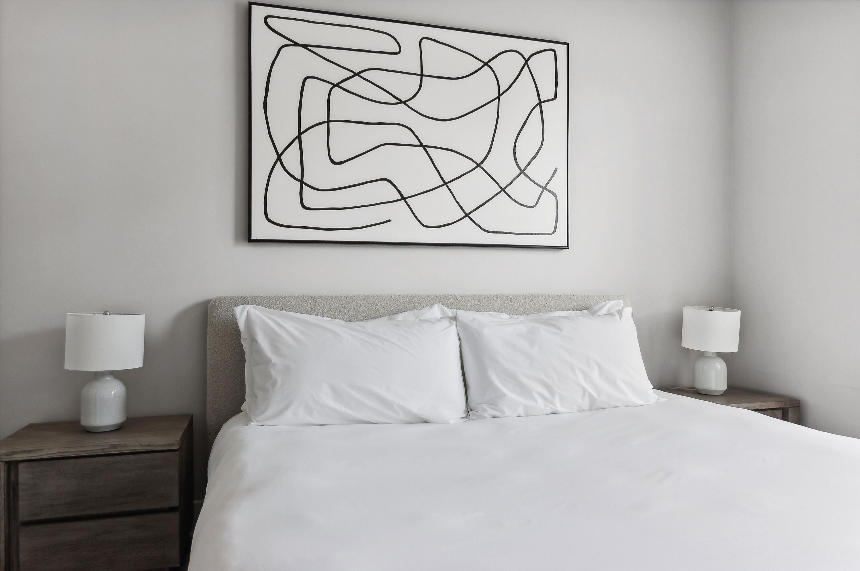 Cama bem arrumada com lençóis e almofadas brancas, um quadro abstrato moderno na parede e duas mesas de cabeceira com abajures.