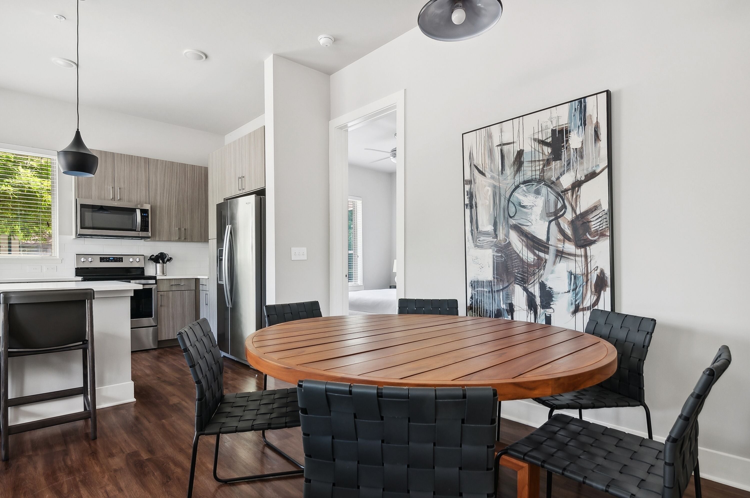 A modern kitchen with a central island, black woven chairs, and a large abstract painting on the wall.