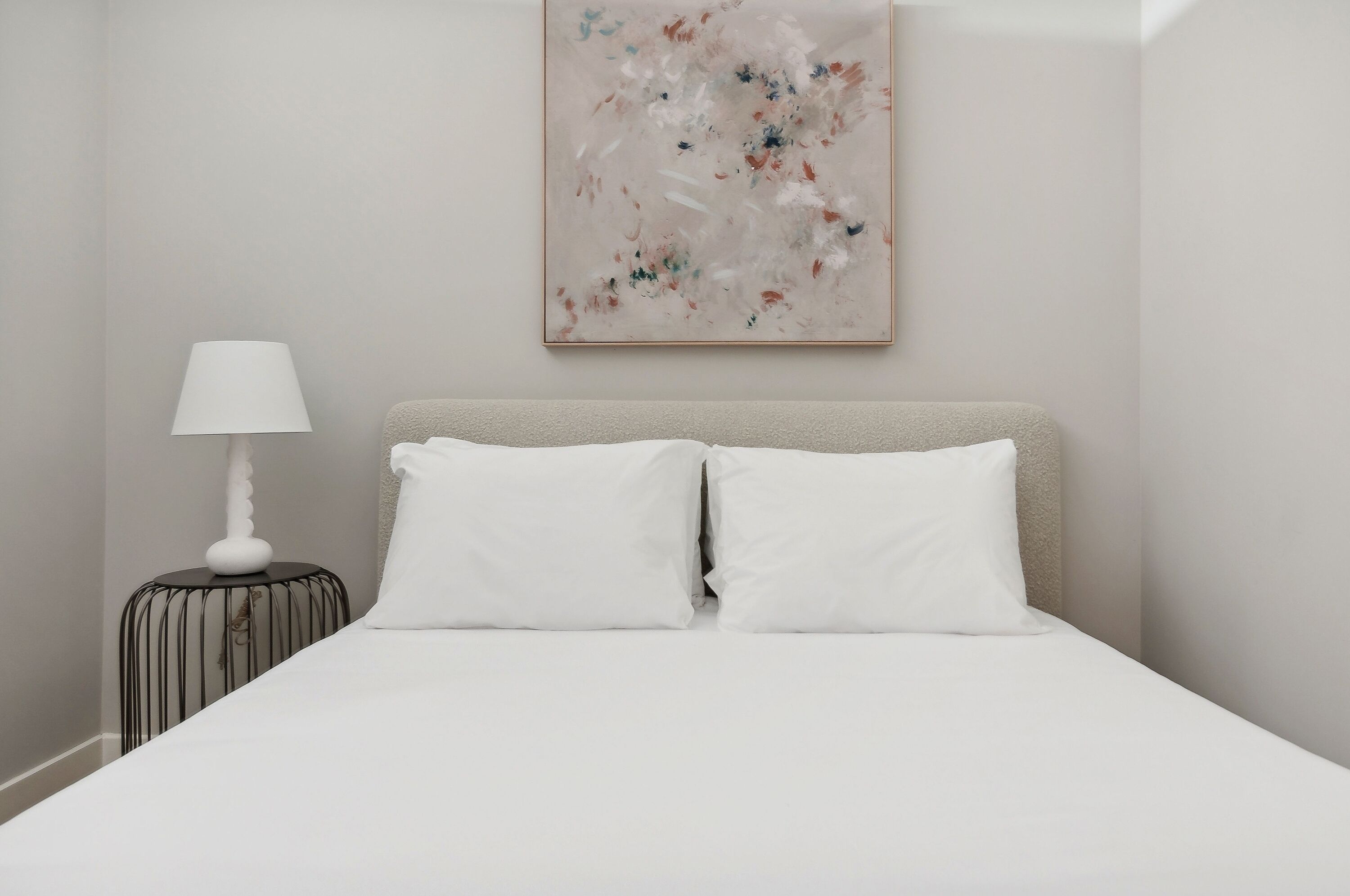 A neatly made bed with white linens and two pillows, a bedside table with a lamp, and a wall-mounted abstract painting.