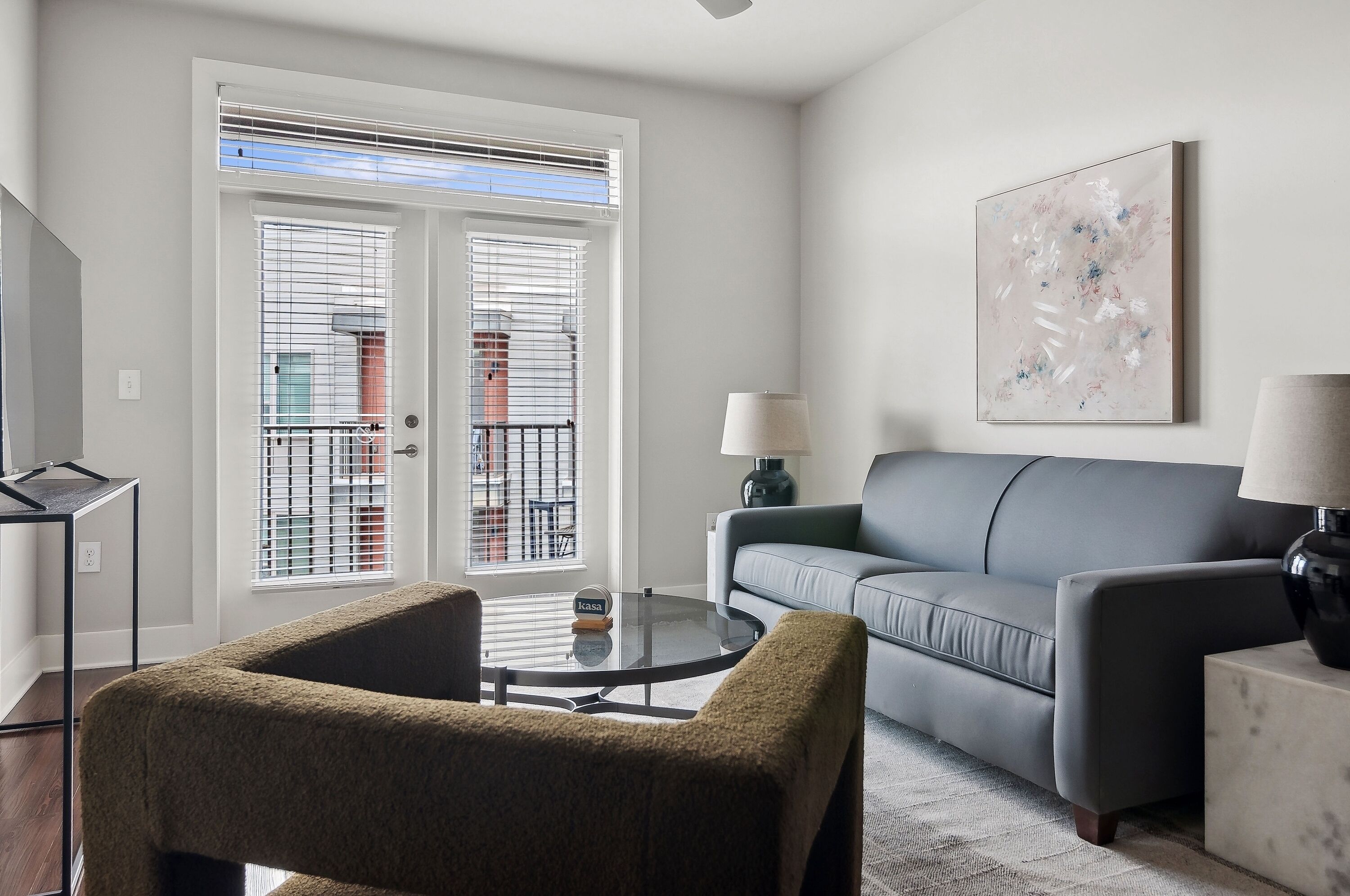 A living room with a grey sofa, a round coffee table, and a small armchair. There is a window with blinds, a lamp, and a painting on the wall.