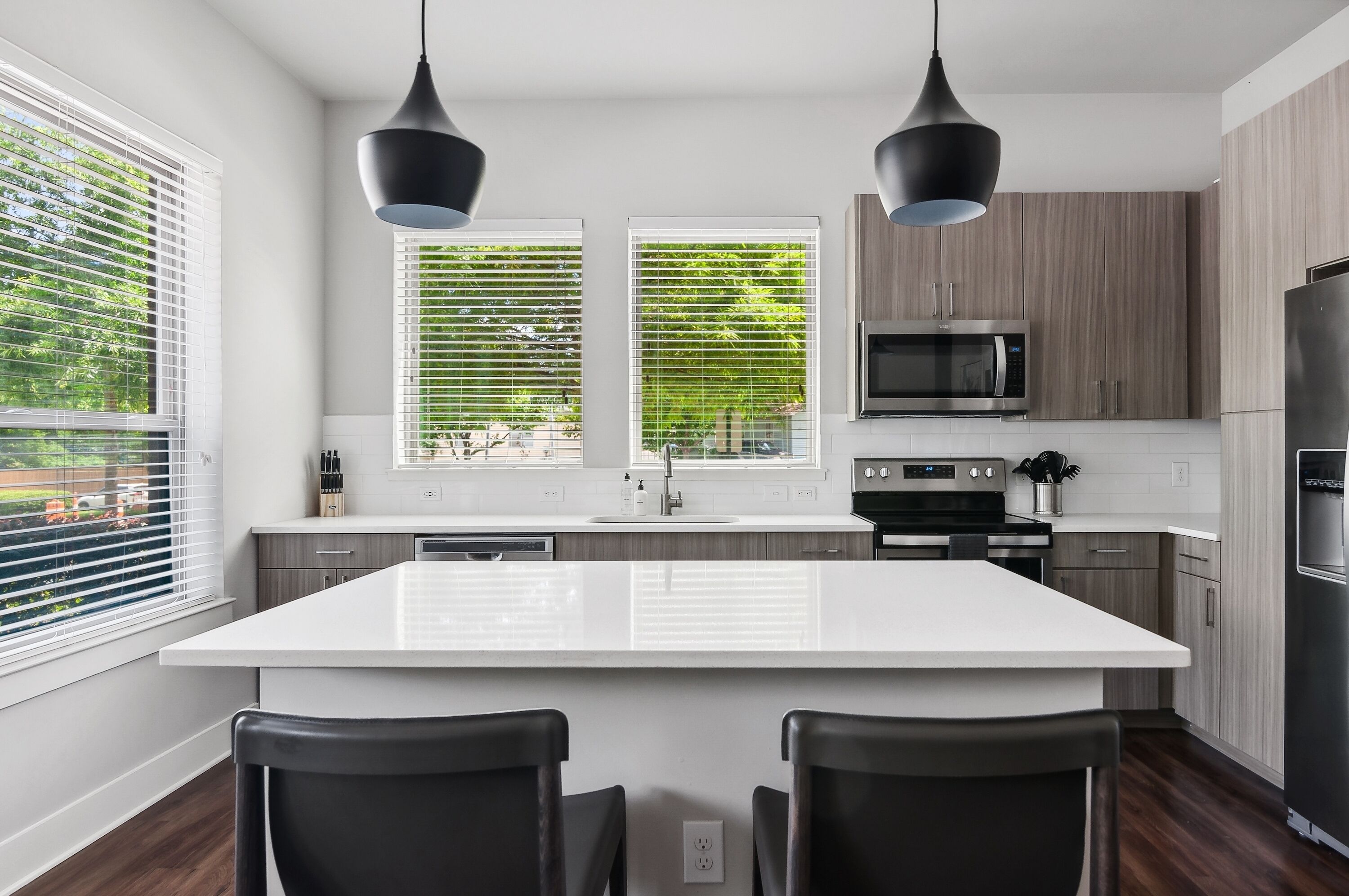 A modern kitchen with a large island, two pendant lights, and a view of greenery outside.