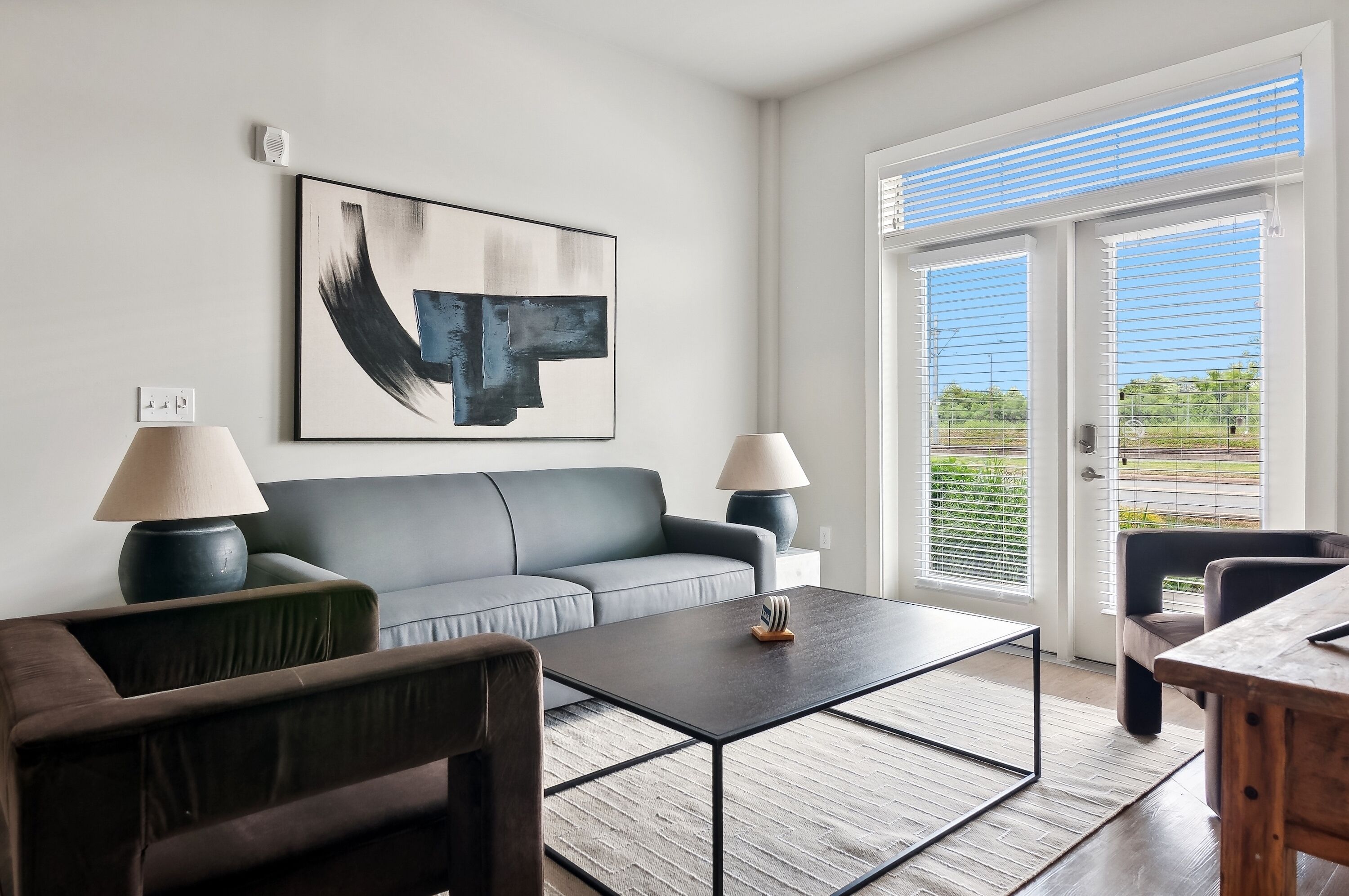 Een moderne woonkamer met een grijze bank, twee bijzettafels met lampen, een salontafel, en een groot abstract schilderij aan de muur.