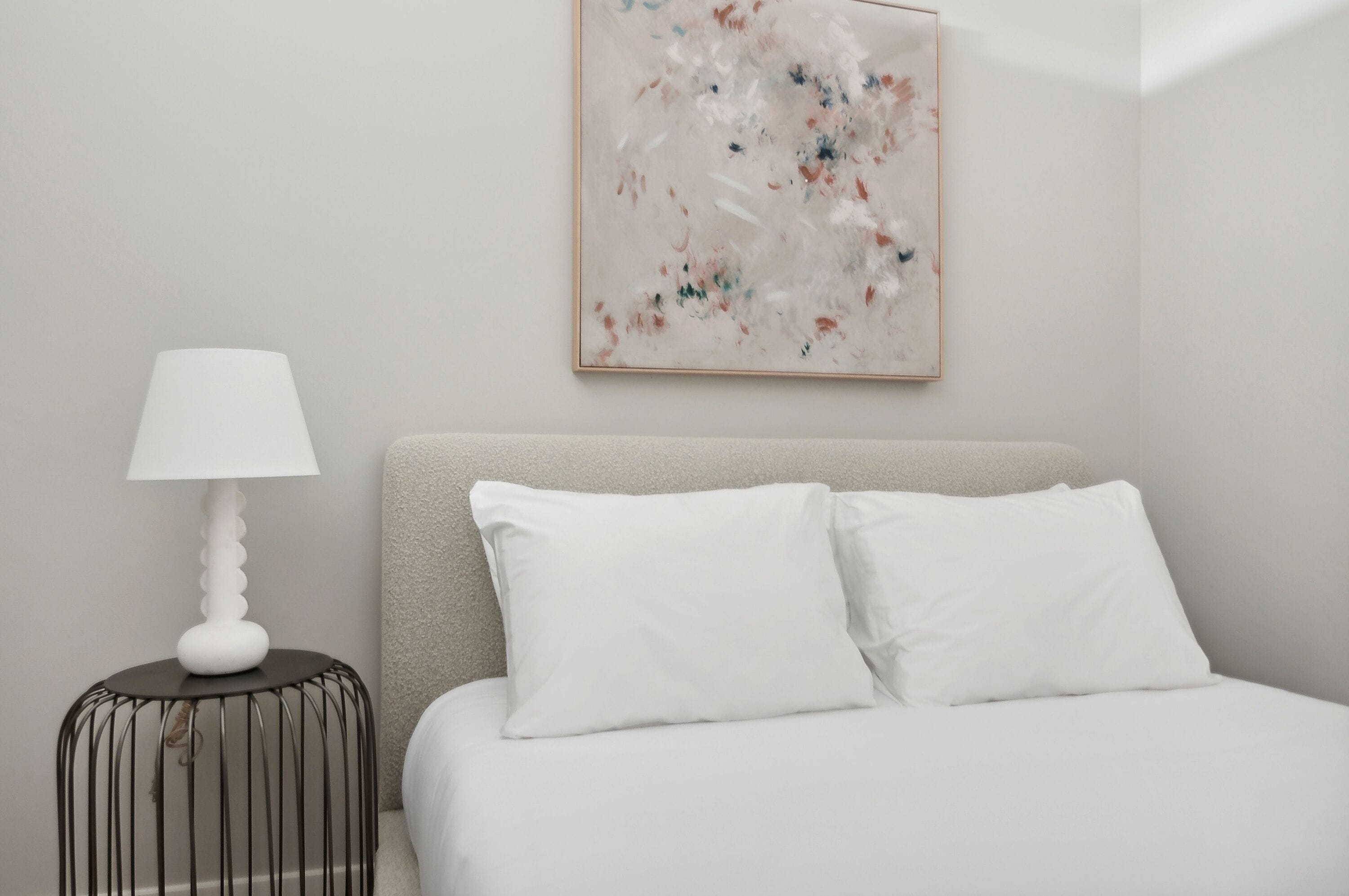 Ein Schlafzimmer mit einem Bett, weißen Kissen, einem Nachttisch mit Lampe und einem Wandbild.
