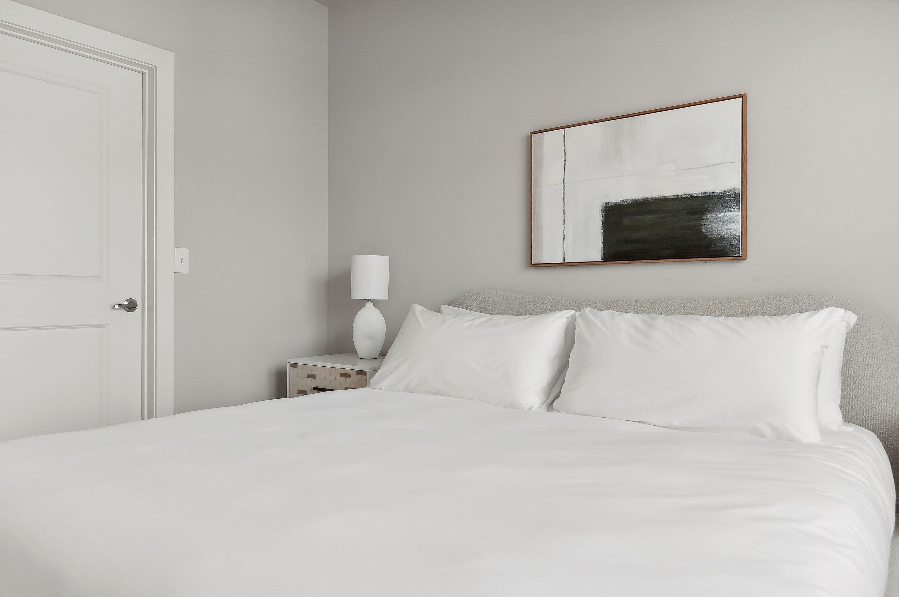 A neatly made bed with white linens, a bedside table with a lamp, and a framed abstract painting on the wall.