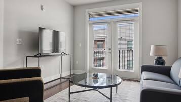 A modern living room with a glass coffee table, a sofa, a TV on a stand, and a balcony with blinds.