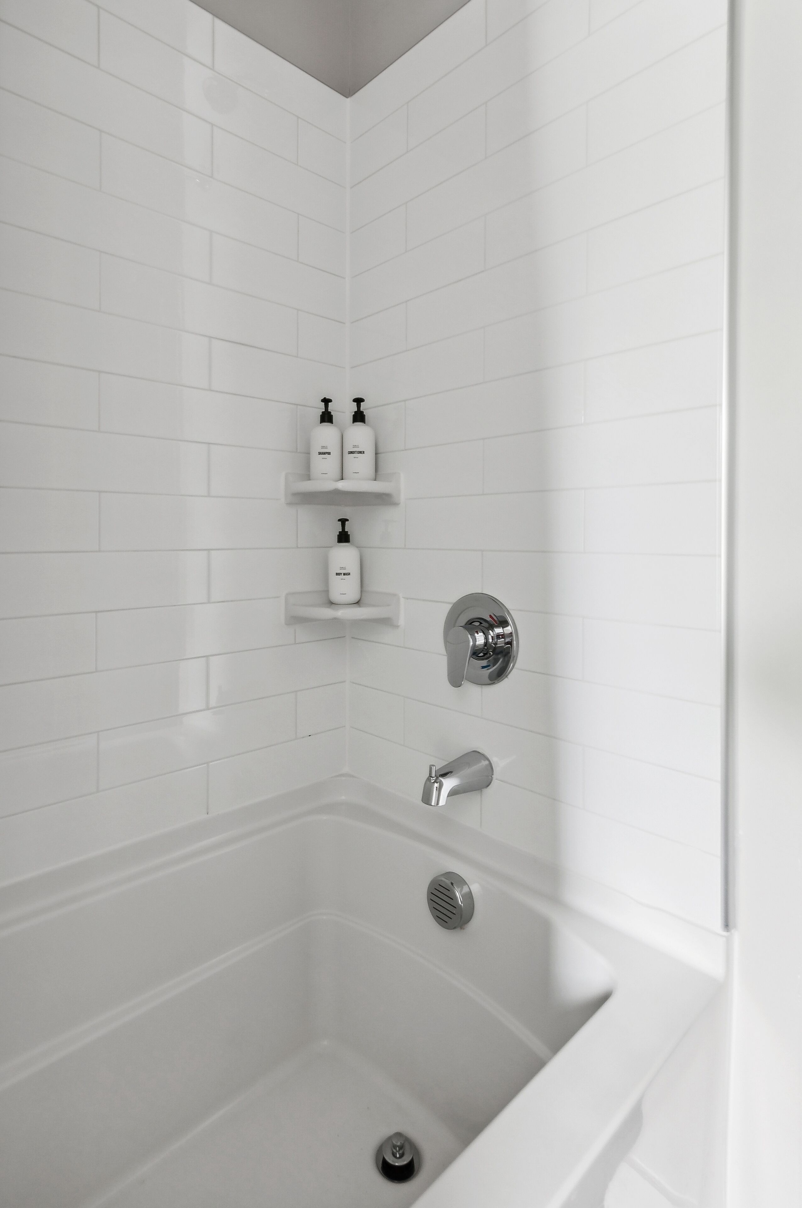 Une salle de bain moderne avec des murs recouverts de carreaux blancs, une baignoire blanche et deux étagères contenant des articles de toilette.
