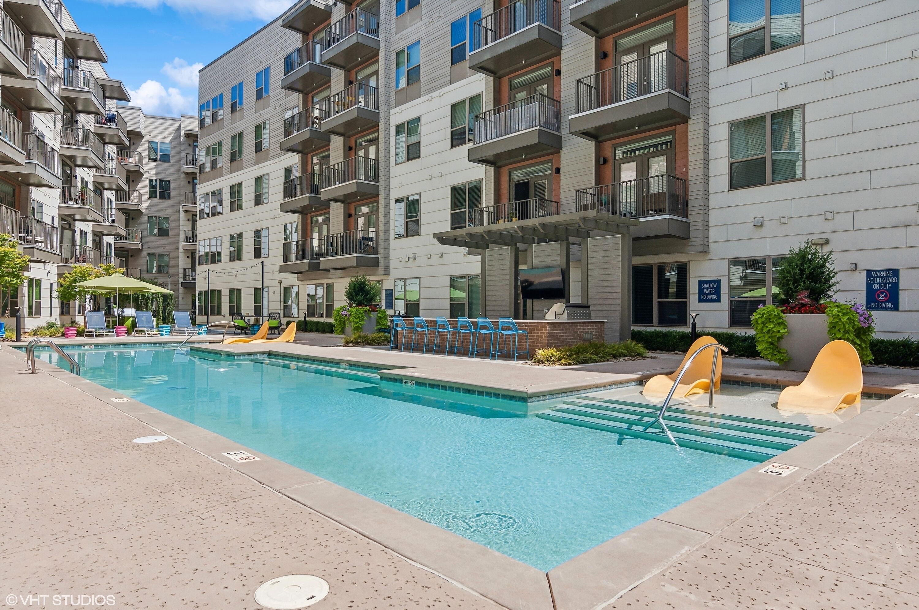 A swimming pool area with a pool, lounge chairs, and a building with balconies.