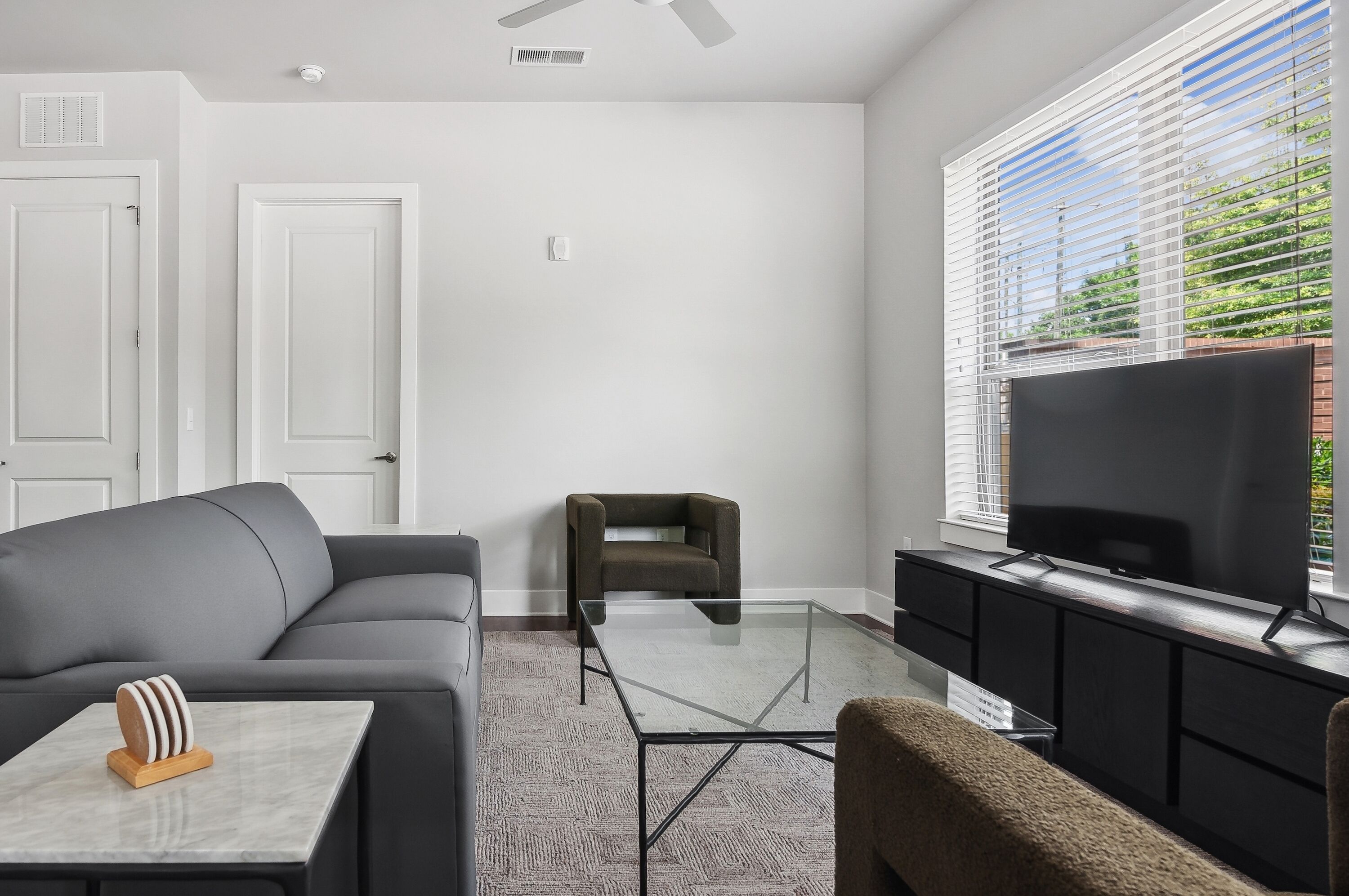A modern living room with a grey sofa, a glass coffee table, a dark armchair, a flat-screen TV on a black stand, and a window with blinds.