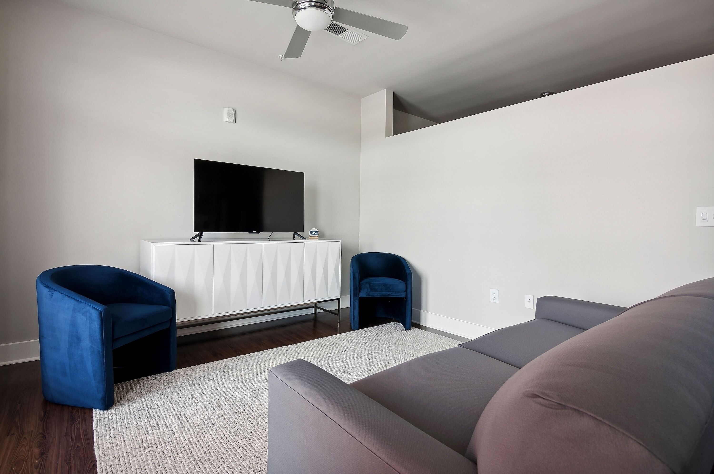 A modern living room with a flat-screen TV mounted on a white cabinet, two blue armchairs, a grey sofa, and a ceiling fan.