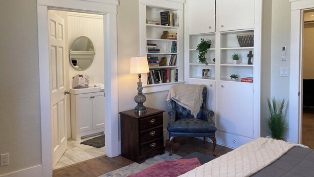 A bedroom with a bed, a chair, a dresser, a lamp, and built-in shelving with books and decorative items.