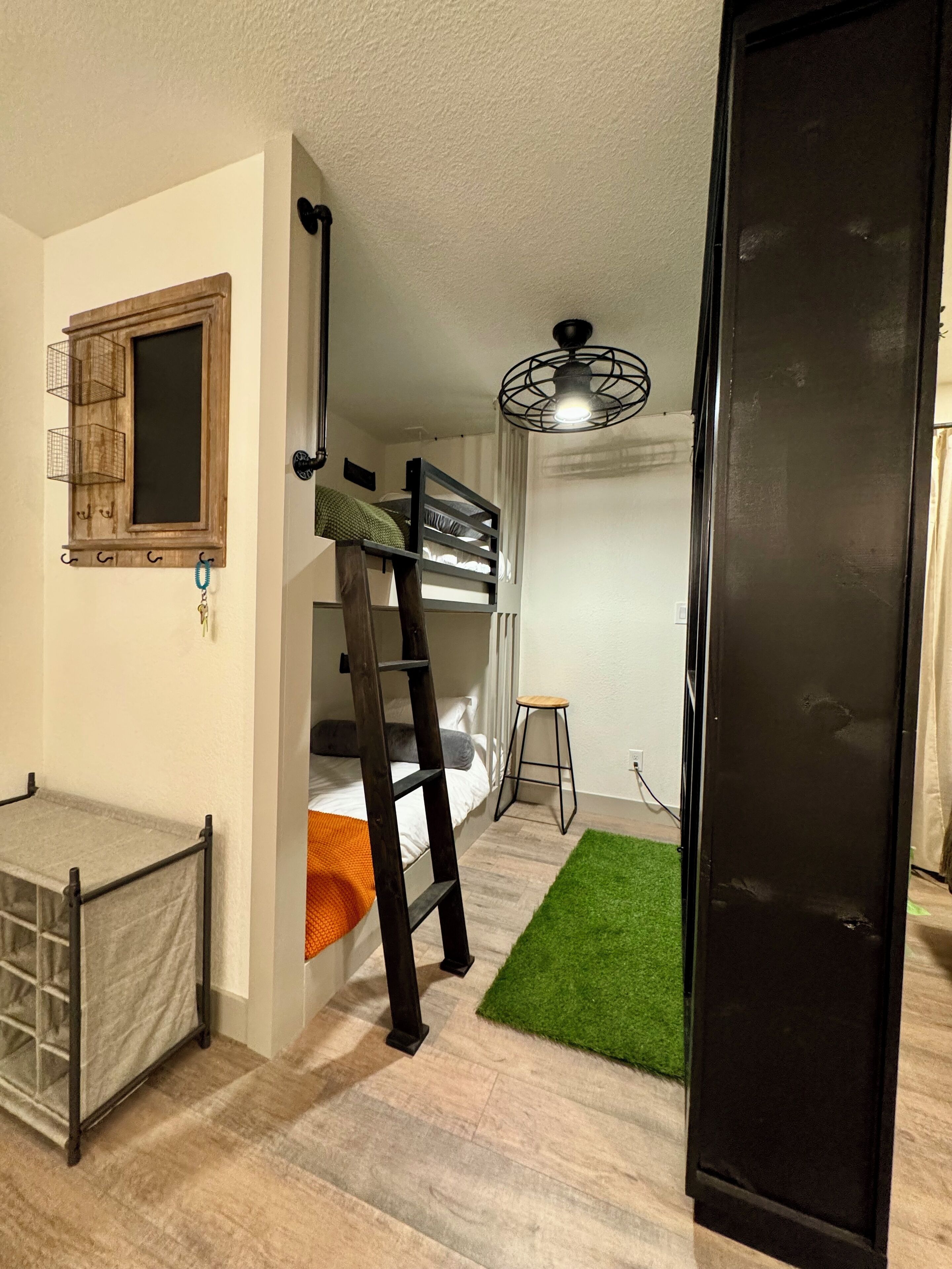 Une petite pièce comprenant un lit superposé, un petit tabouret, un tapis vert et un réfrigérateur noir.