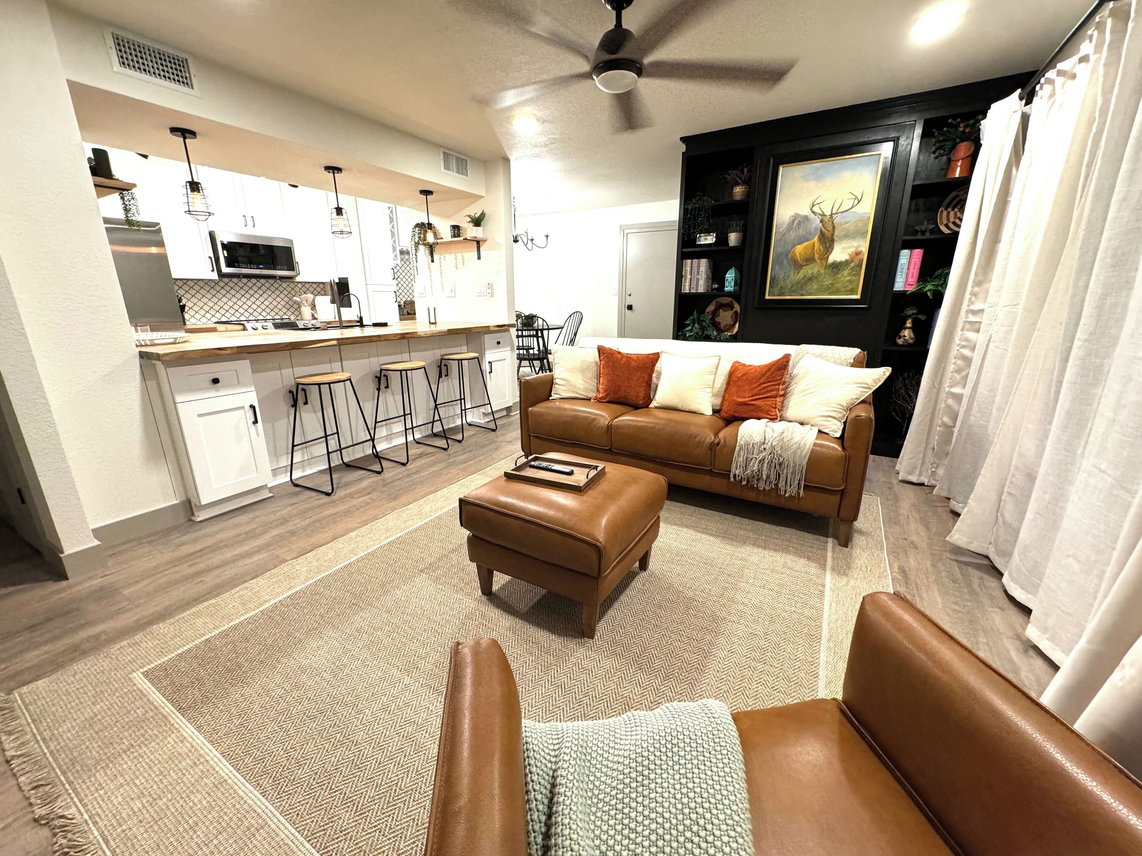 A modern living room with a brown leather sofa, a coffee table, and a kitchen area with white cabinets and a bar stool.