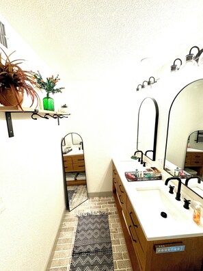 A narrow bathroom with a white sink, a mirror, and a small shelf with a potted plant and a green bottle. - The Cozy Getaway  (Colorado Springs)