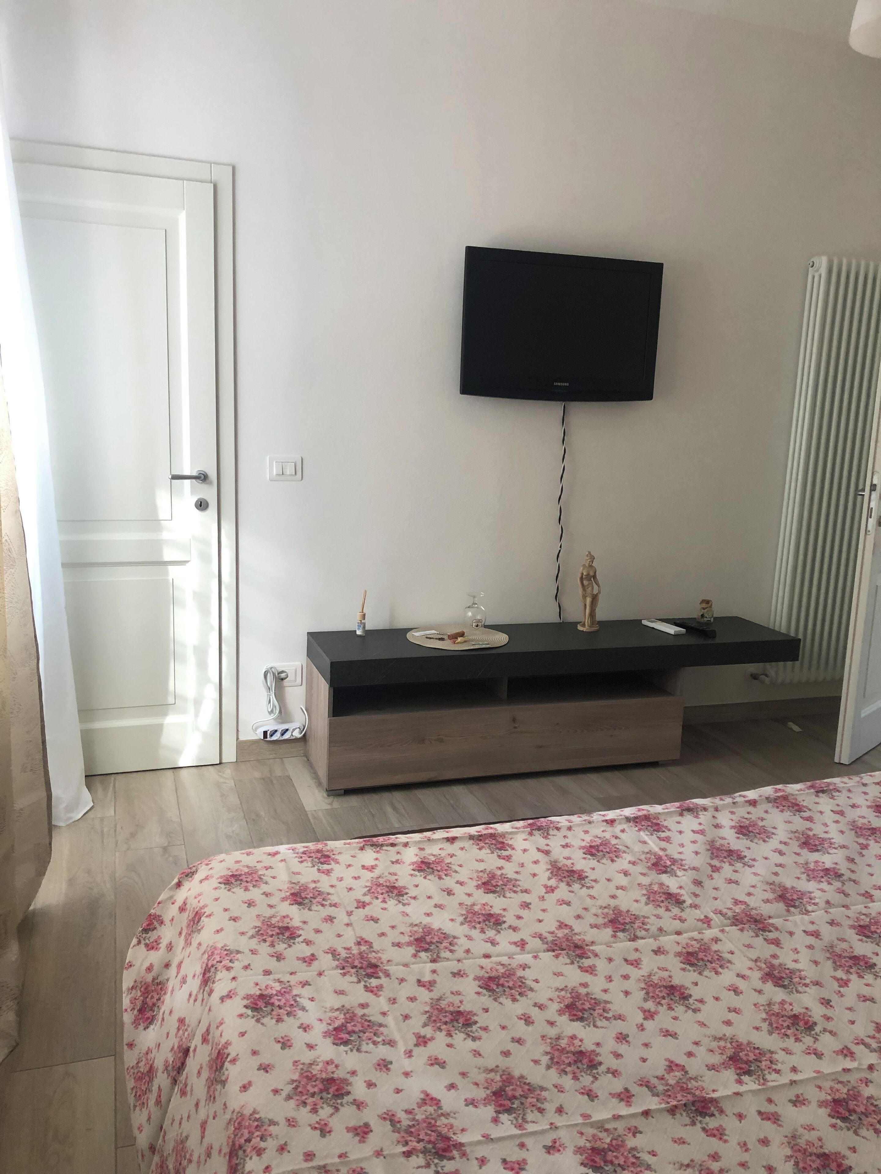 A bedroom with a bed, a TV mounted on the wall, a bedside table with a lamp, and a radiator.