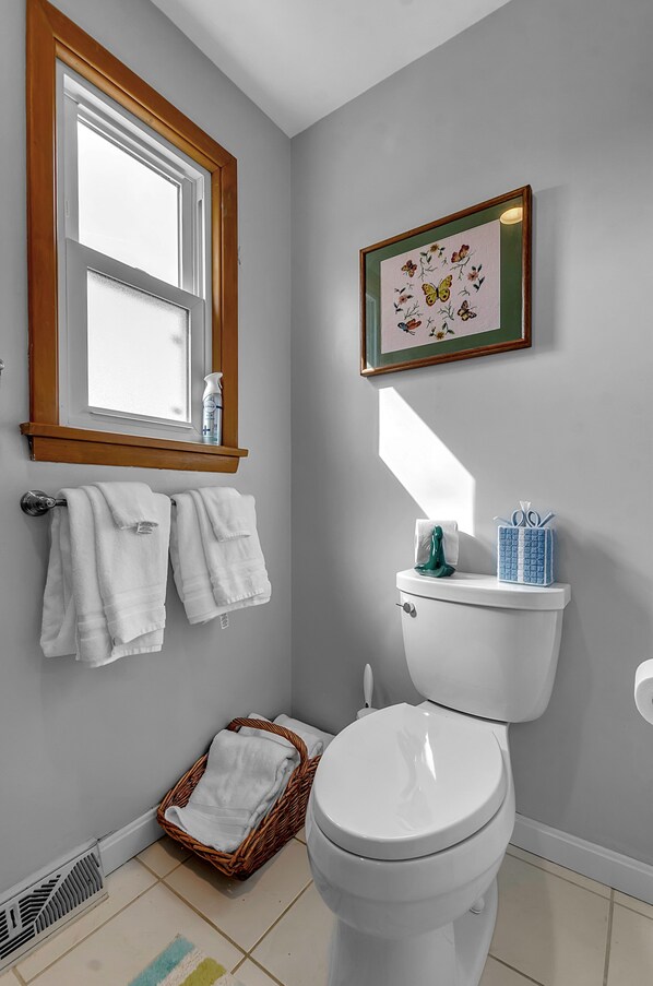 A bathroom with a toilet, a window, a framed floral artwork, and a basket of towels.