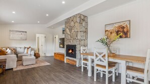 A modern living room with a stone fireplace, white furniture, and a dining area. - Nowhere Cottage rural retreat (Crackenback)