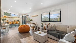 A modern living room with a large window, a cozy sofa, a wooden dining table, and a kitchen in the background. - Nowhere Cottage rural retreat (Crackenback)