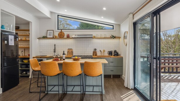 A modern kitchen with a central island, bar stools, a built-in oven, and a large window overlooking a deck. - Nowhere Cottage rural retreat (Crackenback)