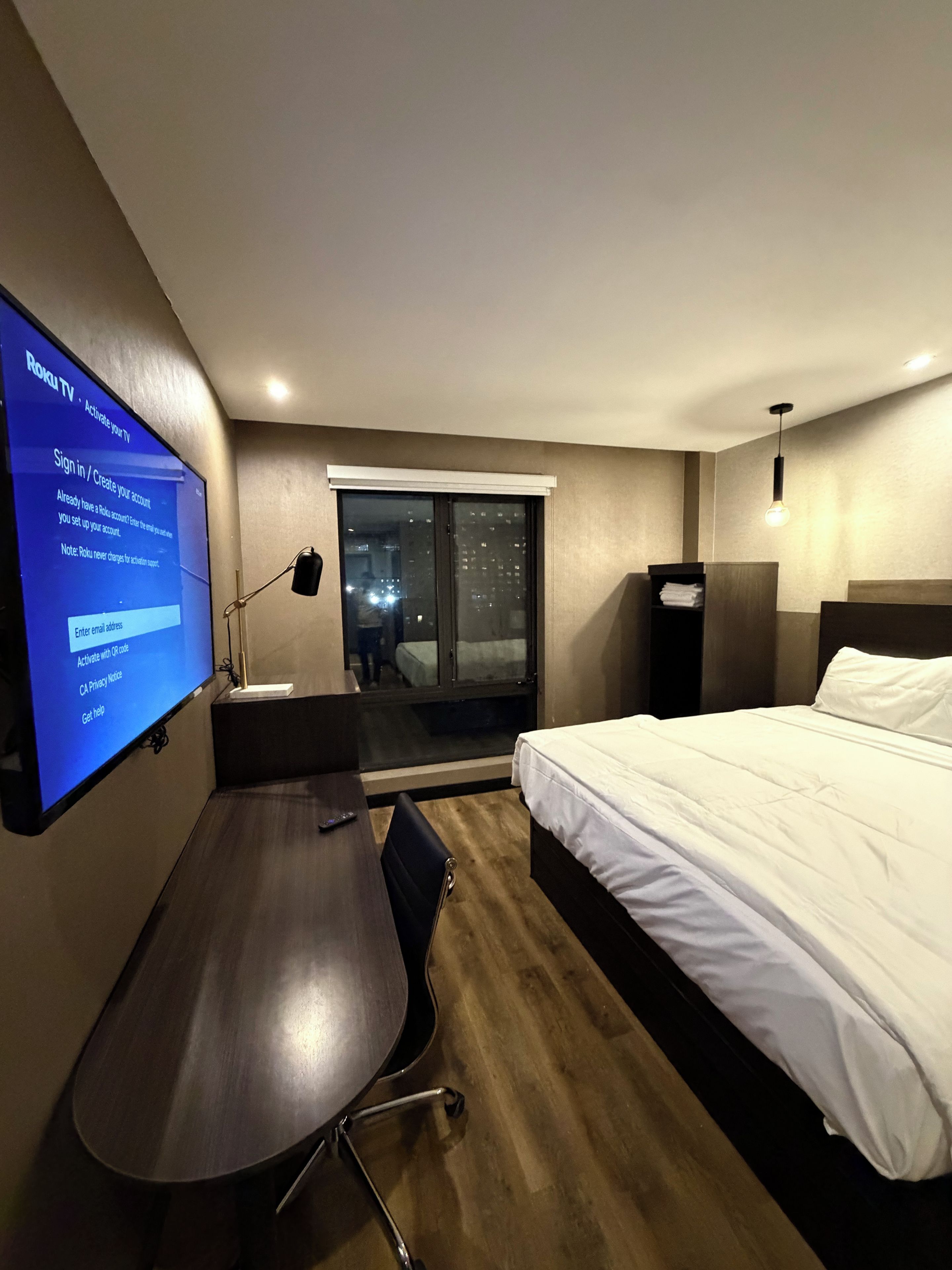 A hotel room with a bed, a desk, a TV, and a view of the city at night.