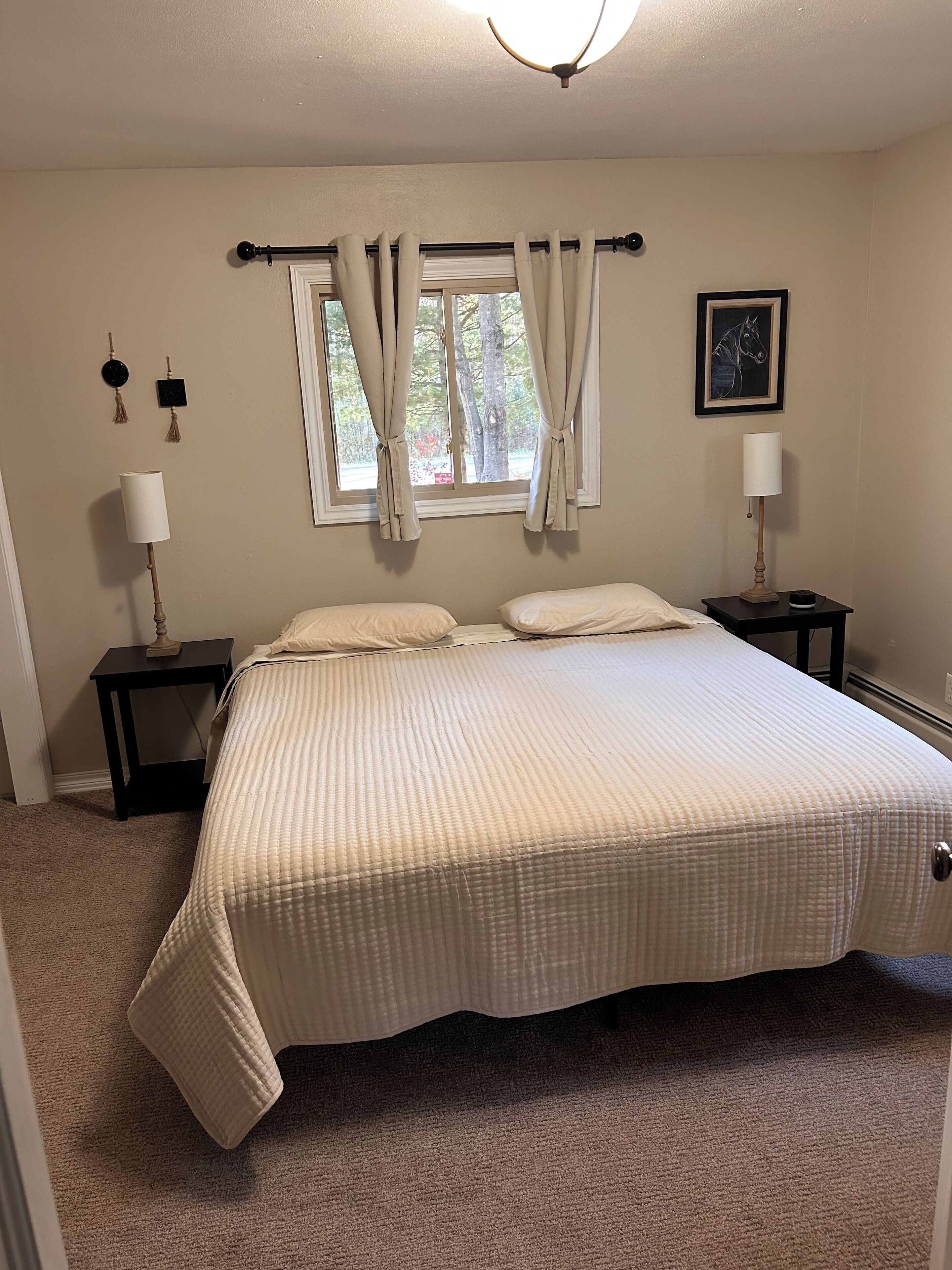 Un dormitorio con una cama grande, dos mesitas de noche, una ventana con cortinas, un ventilador de techo y un cuadro en la pared.
