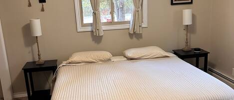 Un dormitorio con una cama grande, dos mesitas de noche, una ventana con cortinas, un ventilador de techo y un cuadro en la pared.