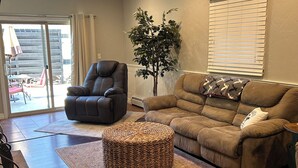 A living room with a brown leather sofa, a dark leather armchair, a woven ottoman, a potted plant, and a large window with blinds. - Close to Everything! Freshly updated ranch home. (Shawano)