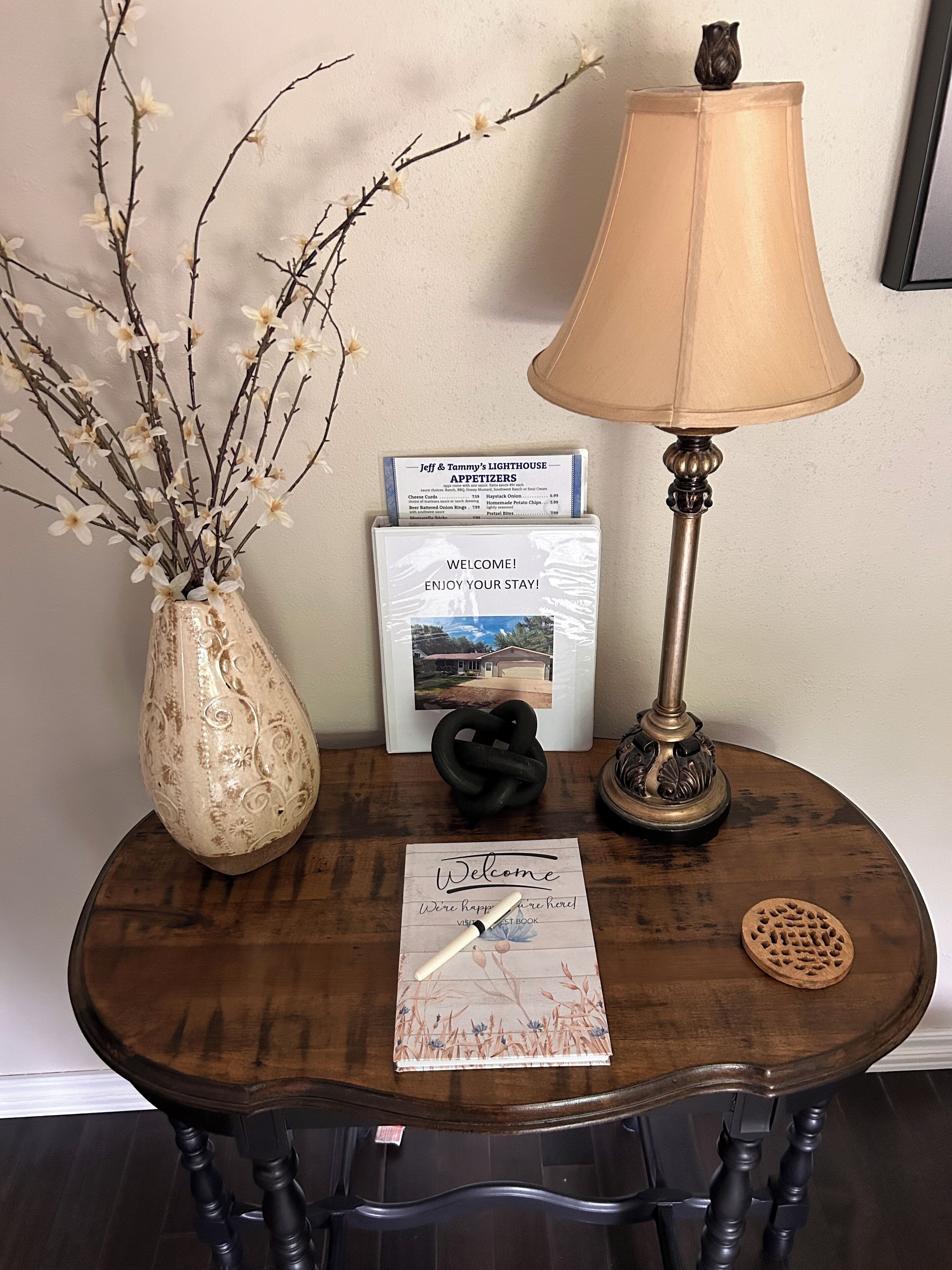 Una mesa de madera con un jarrón de flores blancas, una lámpara, un cartel de bienvenida, un menú y un cuadro enmarcado.