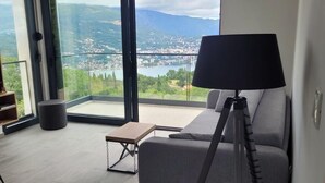 A modern living room with a sofa, a small table, and a tripod lamp. Large windows offer a view of a mountainous landscape.