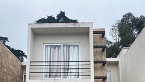 Een modern gebouw met twee verdiepingen, een plat dak, een balkon met een metalen hekwerk en een kleine tuin met een pad naar de ingang.