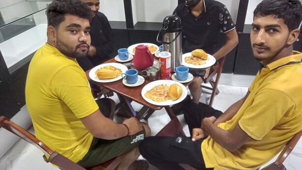 Un groupe de quatre personnes est assis autour d’une table, devant des assiettes remplies de nourriture, des tasses et une bouilloire.
