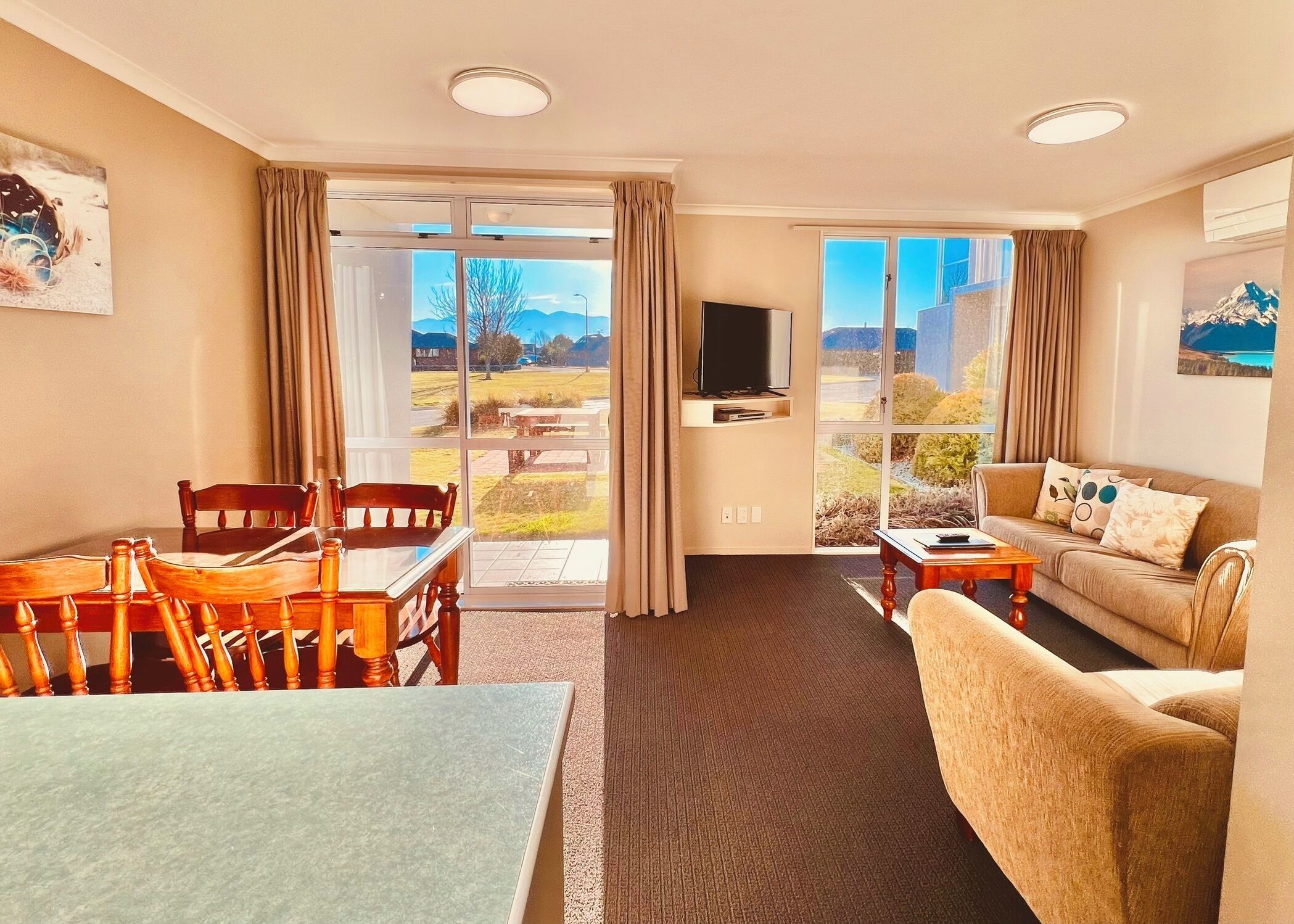 A hotel room with a dining table, chairs, a sofa, a TV, and large windows offering a view of the outdoors.