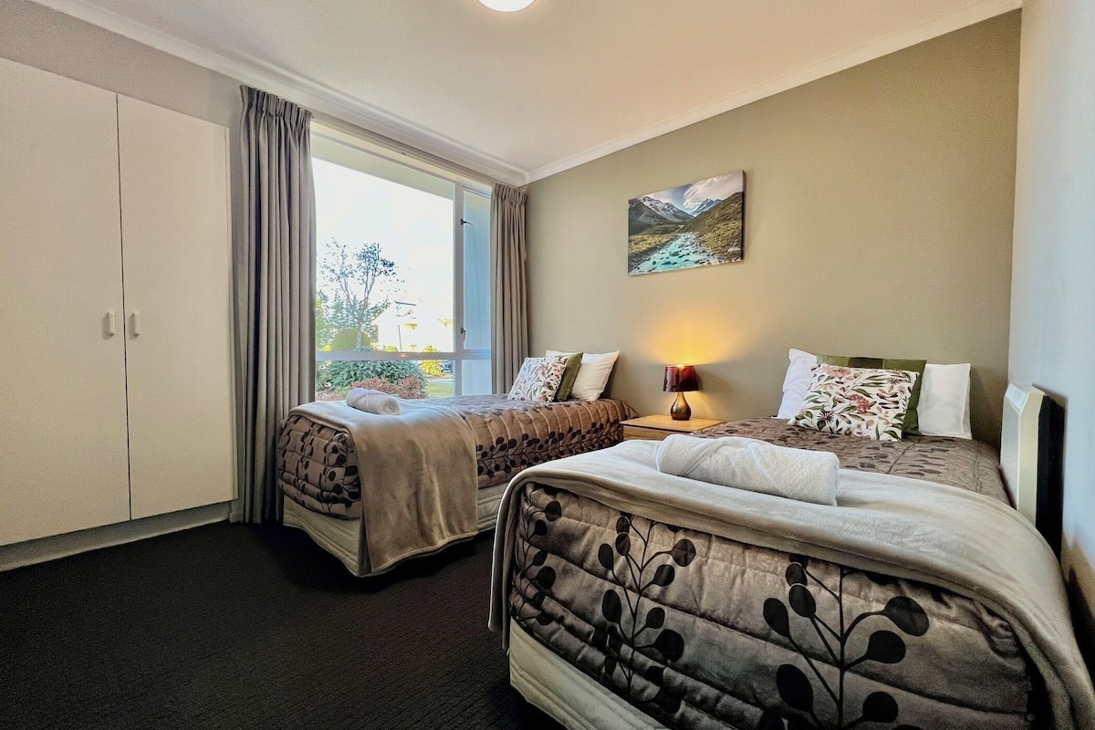 Habitación de hotel con dos camas, un ventanal grande, un cuadro y una lámpara.