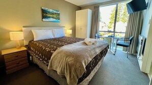 A hotel room with a bed, bedside tables, a desk, a chair, a TV, and a window with curtains. - Brinkley Resort (Methven)