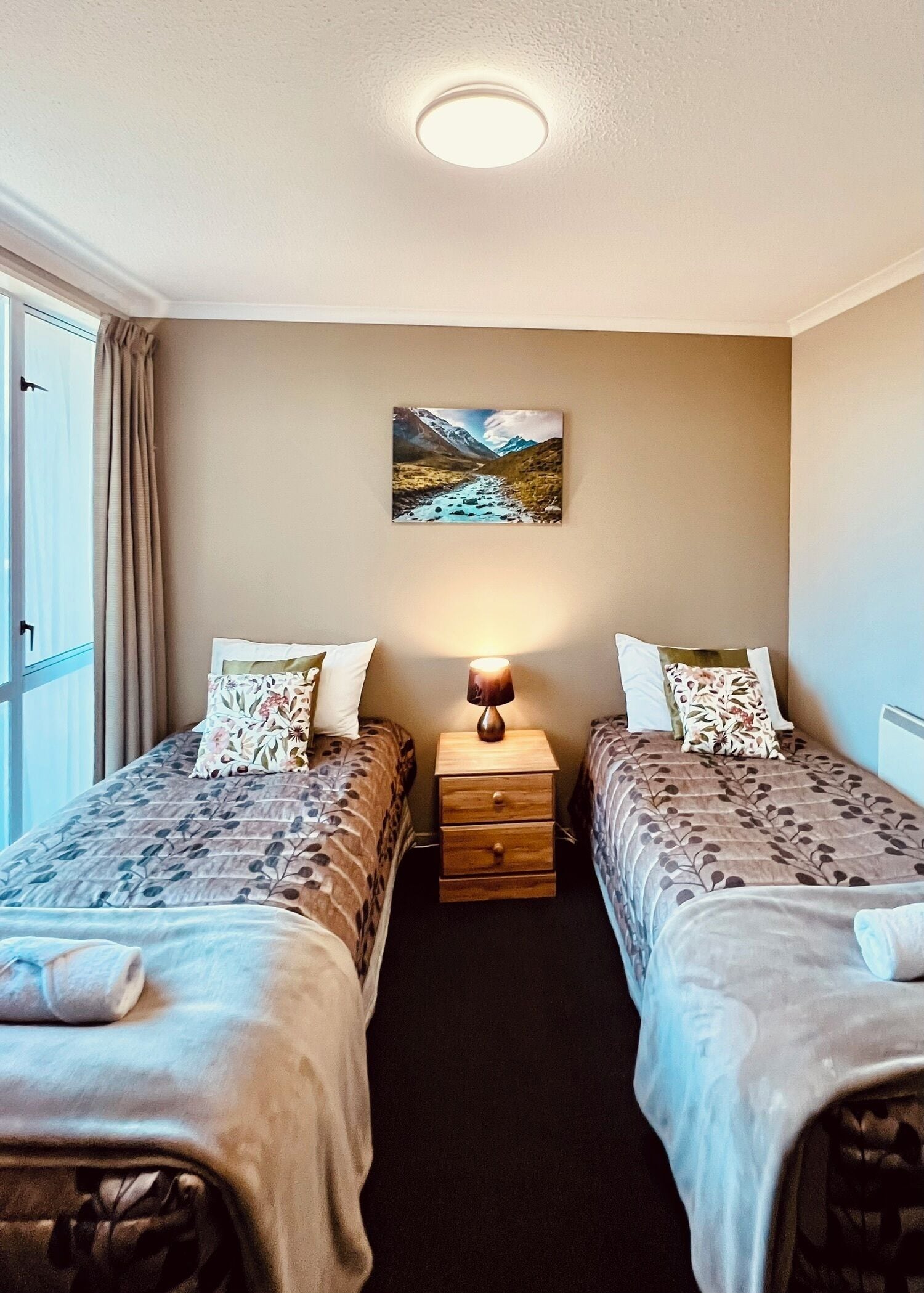 Una habitación con dos camas, una mesita de noche con una lámpara, un cuadro en la pared y una ventana con cortinas.