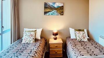 Una habitación con dos camas, una mesita de noche con una lámpara, un cuadro en la pared y una ventana con cortinas.