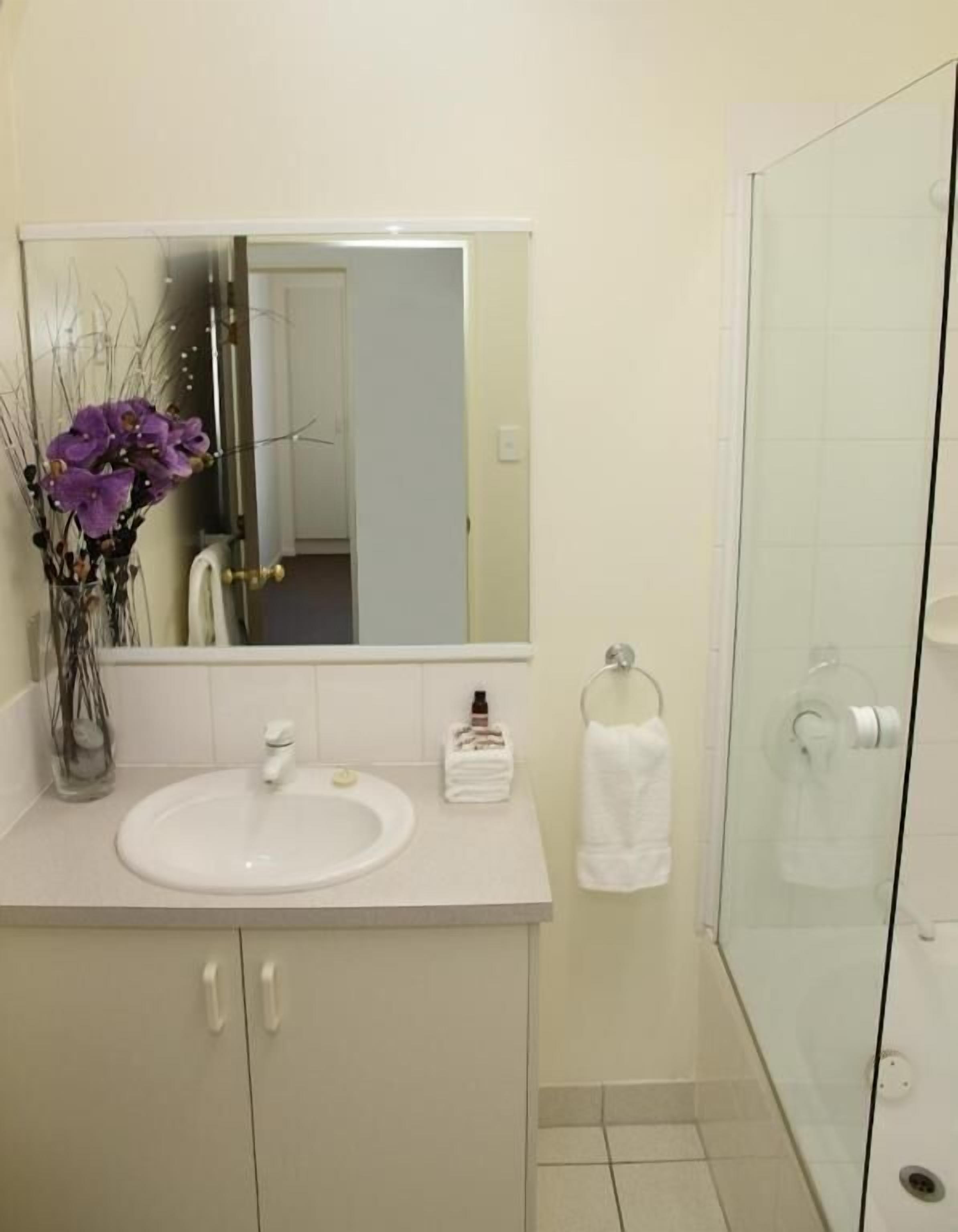 A bathroom with a white sink, a mirror, a glass shower enclosure, and a vase with purple flowers.