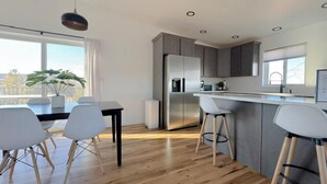 A modern kitchen with a central island, bar stools, a dining table with chairs, and a large window with a view.