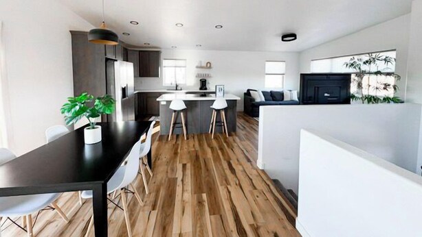 A modern kitchen with a central island, a dining area with a black table and white chairs, and a living area with a sofa and a TV.