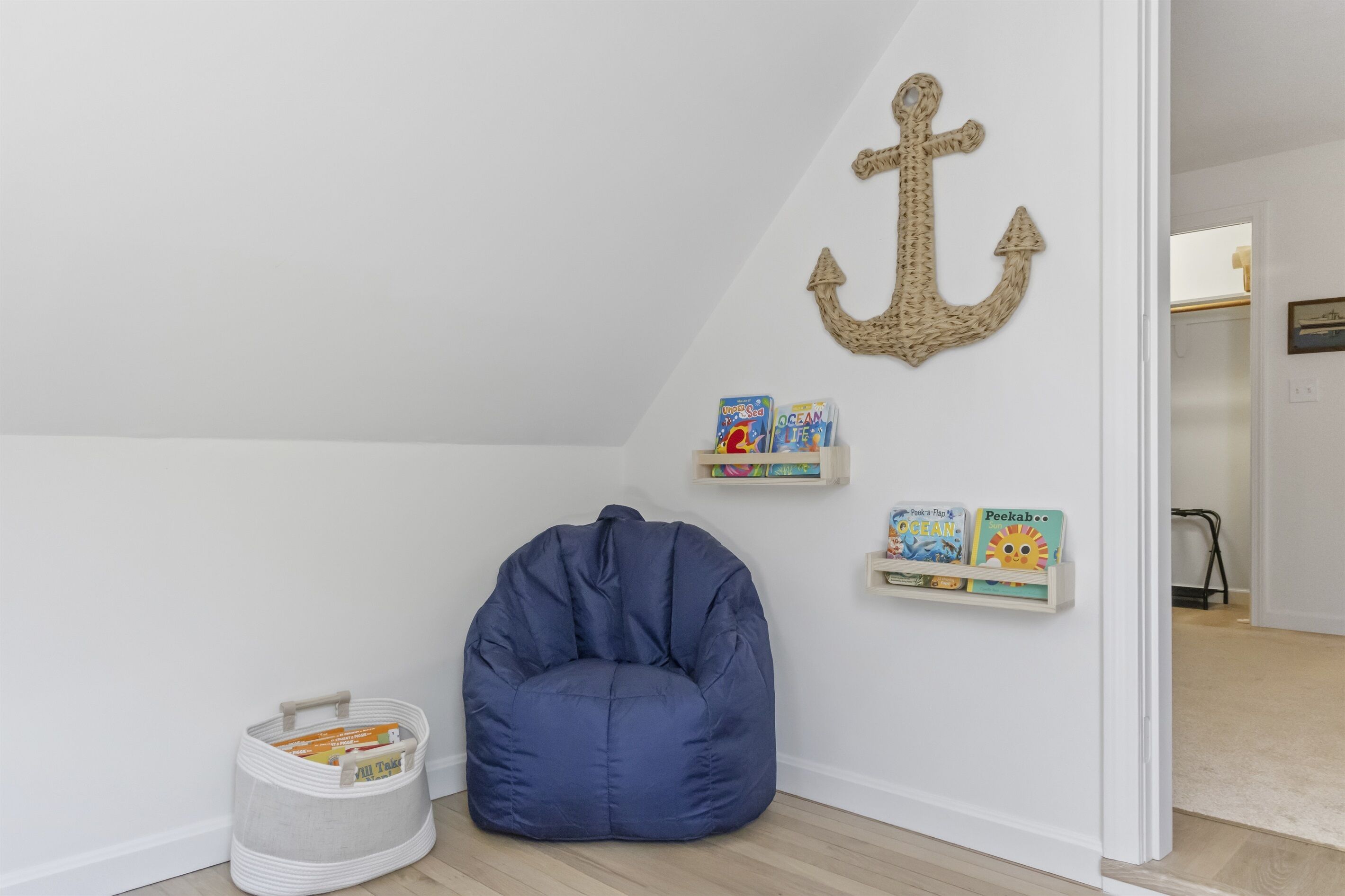 Una habitación con un sillón de peluche azul, una cesta blanca y una pared decorada con un ancla y libros infantiles.