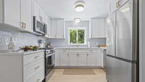 A modern kitchen with white cabinetry, stainless steel appliances, and a window providing natural light. - Trendy Great Pond Escape! HotTub Open! Sleeps 9! (Falmouth)