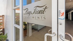 L’entrée des appartements Bay Front se distingue par une porte en verre transparent, une rampe métallique et une enseigne indiquant qu’il faut tirer pour ouvrir.