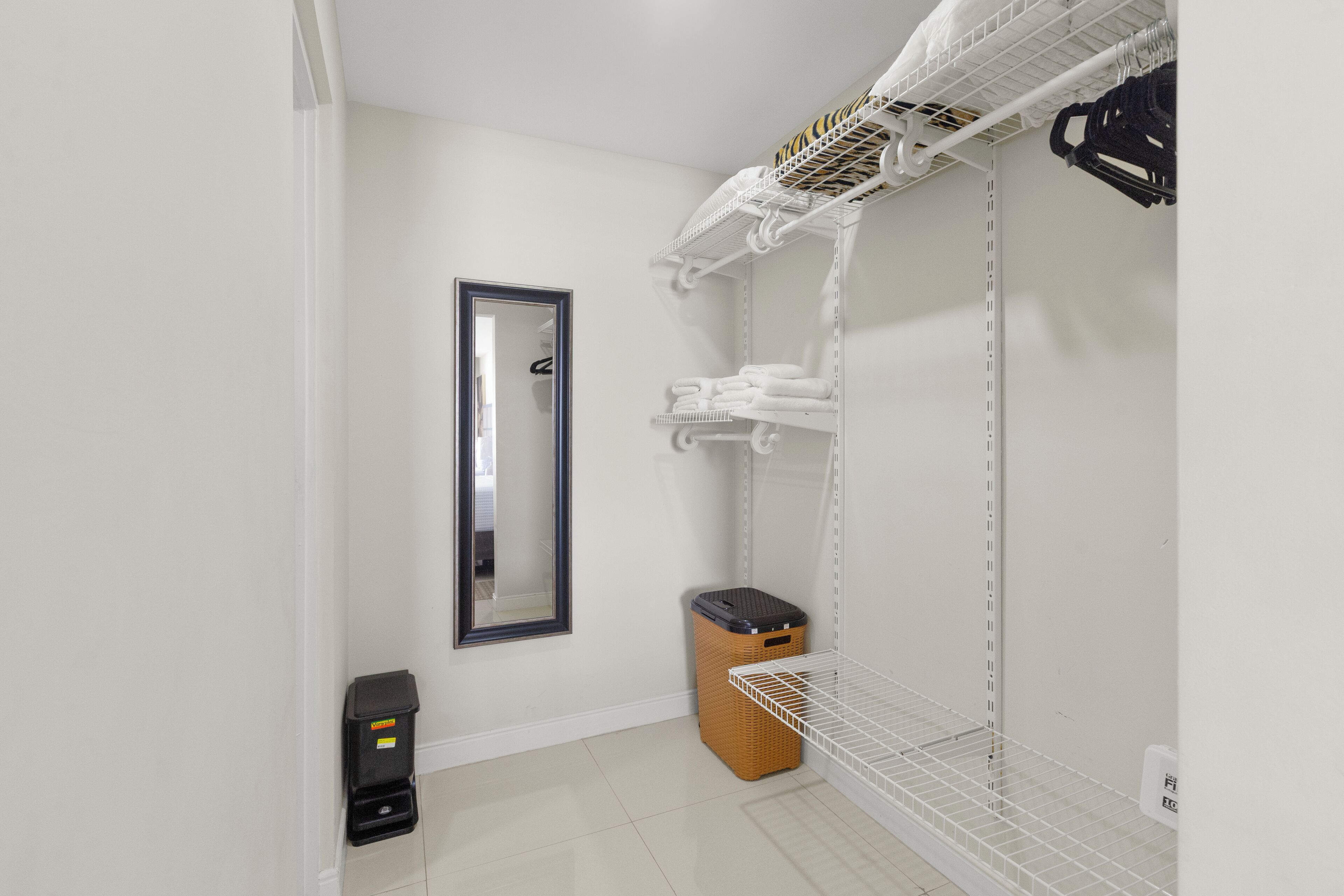 Un baño limpio y ordenado con un espejo, una repisa con toallas, un cubo de basura y una cesta de ropa.