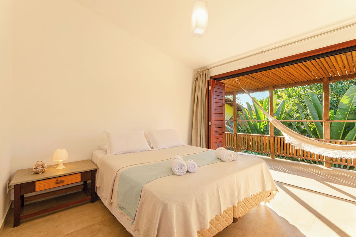 Un dormitorio con cama, mesita de noche, lámpara, hamaca y vistas al verdor exterior.