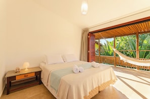 A bedroom with a bed, a nightstand, a lamp, a hammock, and a view of greenery outside. - Pousada Vila Gostoso (São Miguel do Gostoso)