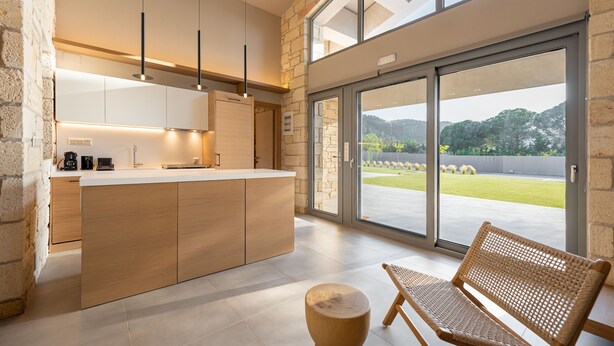 Una cocina moderna con armarios de madera, una isla central, luces colgantes y un amplio ventanal corredera que da a un patio con vistas a la vegetación.