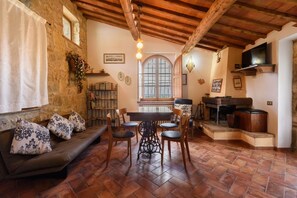 A rustic interior with stone walls, a wooden ceiling, a dining area with a table and chairs, and a sofa with patterned cushions.