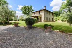 A stone house with a gravel driveway, surrounded by greenery and trees.