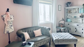 A living room with a grey sofa, a matching ottoman, a vintage radio, and a white cabinet with decorative items. - Audrey- an ode to an icon (Salem)