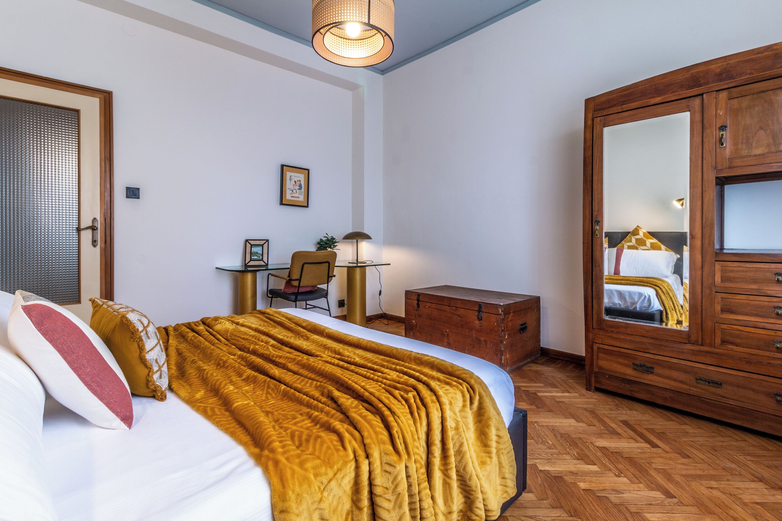 A bedroom with a bed, a wooden wardrobe, a desk with a chair, and a lamp.