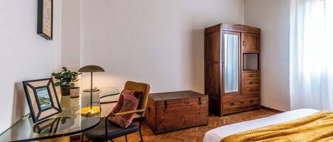 A room with a glass table, a chair, a wooden cabinet, and a bed.